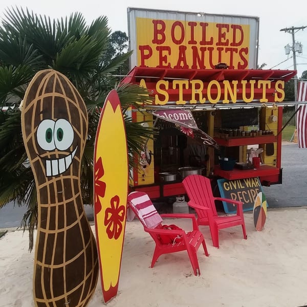 A Boiled Peanuts food truck with a peanut cutout, surfboard, and pink chairs on sand.