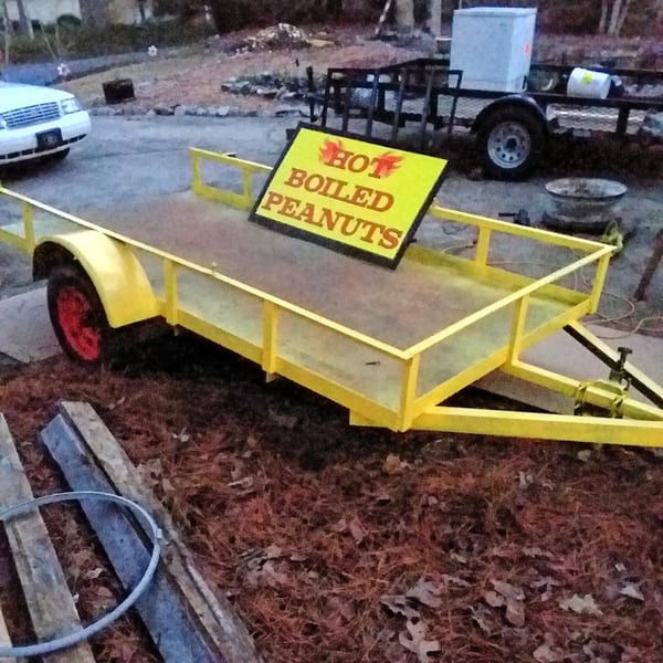Yellow trailer with a "Hot Boiled Peanuts" sign, parked on dirt with a white car in the background.