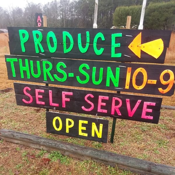Hand-painted wooden sign for "PRODUCE THURS-SUN 10-9 SELF SERVE OPEN" with a yellow arrow.