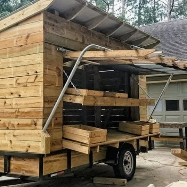 A wooden food cart with shelves and boxes, attached to a trailer, parked outside a garage.