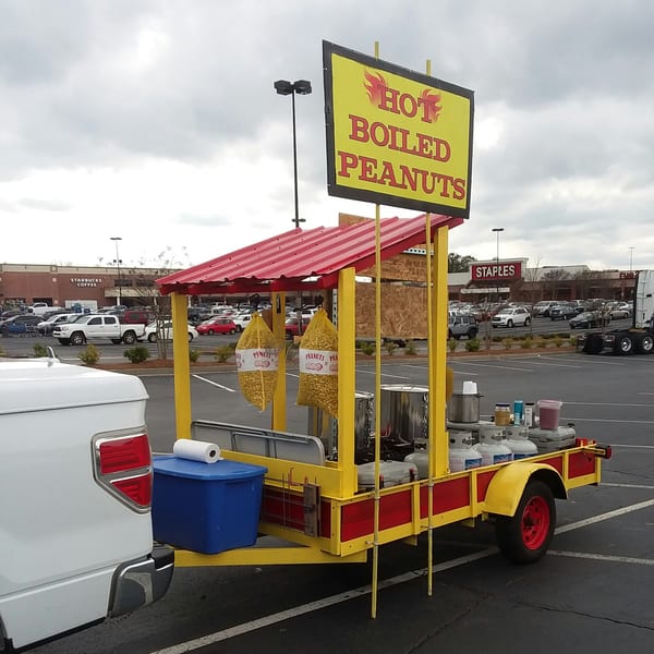 A yellow and red "Hot Boiled Peanuts" stand with two bags of peanuts, parked in a lot.