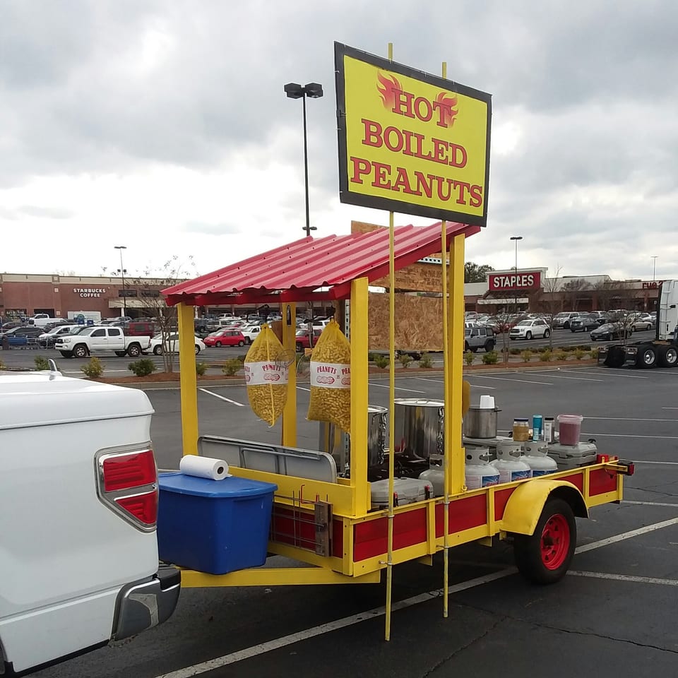 A yellow and red "Hot Boiled Peanuts" stand with two bags of peanuts, parked in a lot.