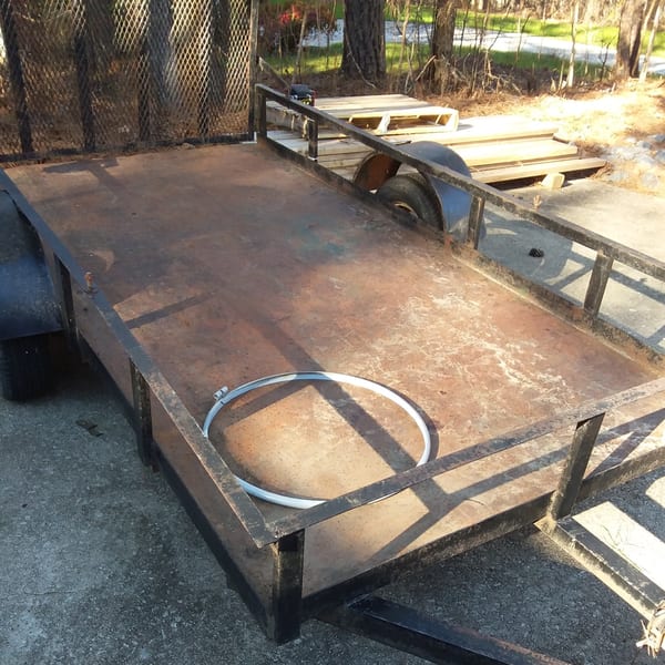 Empty, rusty utility trailer with a metal ring on its bed, parked on concrete near trees.