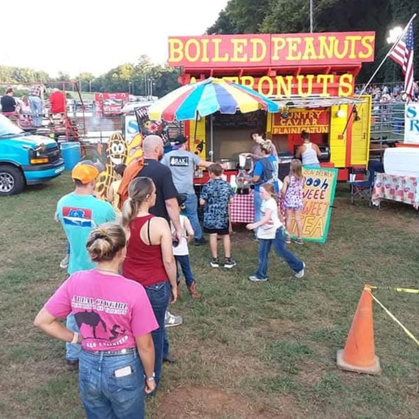People gather around a bright yellow and red "Boiled Peanuts" food stand at an outdoor event.