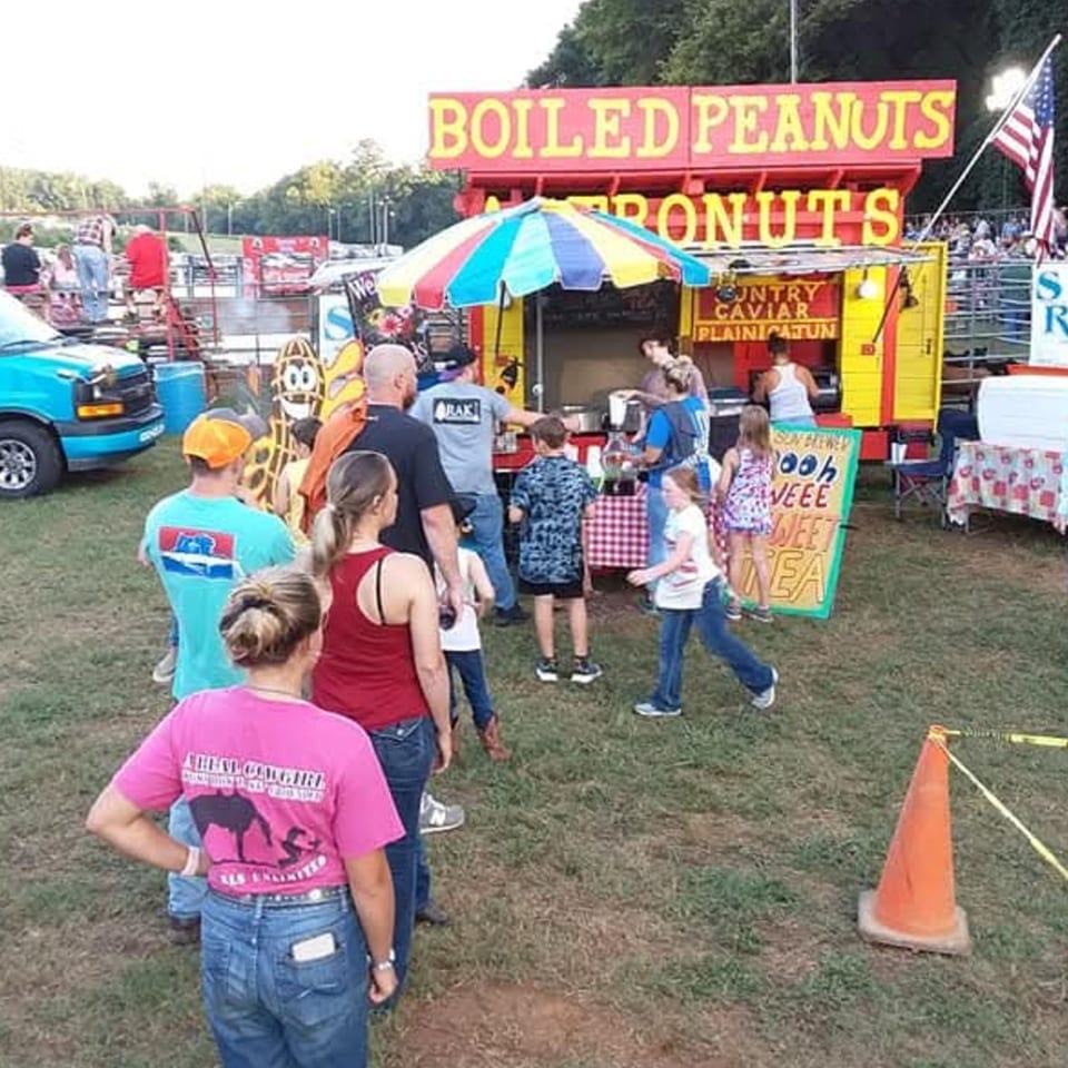 People gather around a bright yellow and red "Boiled Peanuts" food stand at an outdoor event.