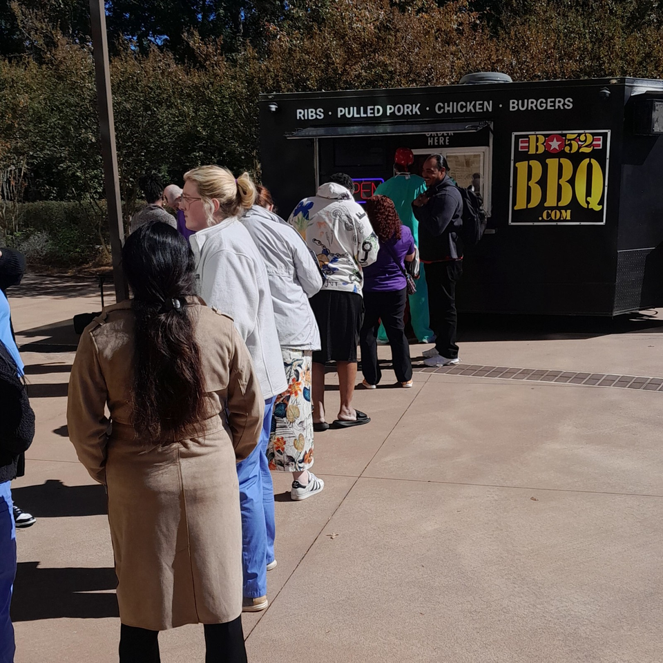Customers queue at a BBQ food truck advertising ribs, pulled pork, chicken, burgers, and BBQ specialties.