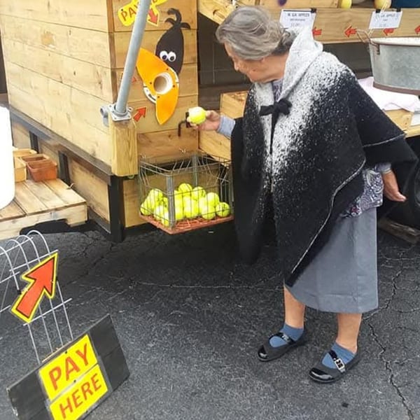 An older woman in a black and white poncho selects a tennis ball from a wire basket at a self-serve stand.