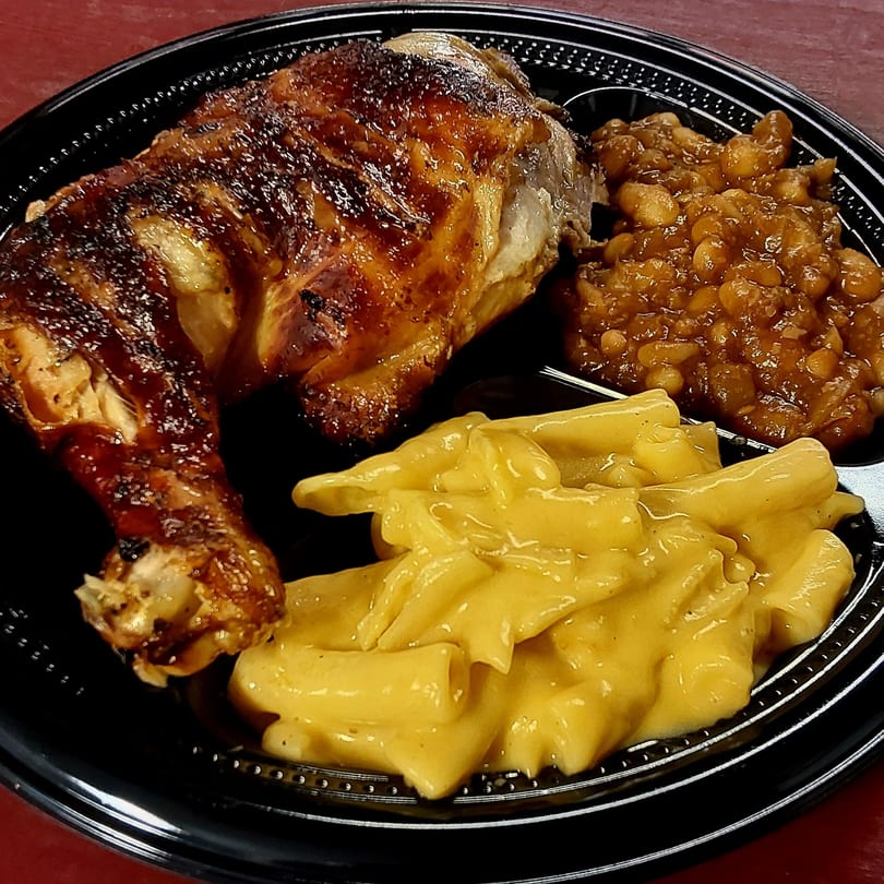 A black plate with a roasted chicken leg quarter, baked beans, and macaroni and cheese.