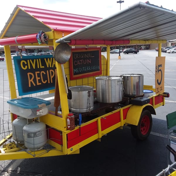 A yellow and red food cart with large cooking pots, propane tanks, and signs for "Cajun Mediterranean" food.
