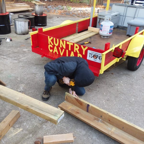 Person drilling wood next to a red and yellow trailer with "KUNTRY CAVIAR" painted on its side.