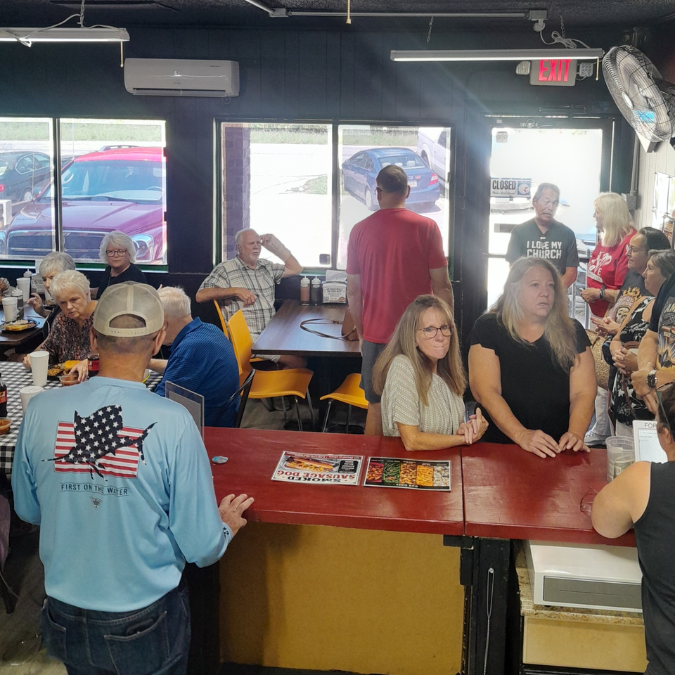Busy casual restaurant interior with diners gathered around tables, colorful chairs, menu cards, large windows, and an exit door.