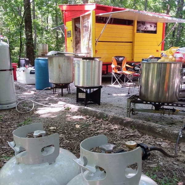 Outdoor cooking setup with large pots on burners, propane tanks, and a yellow food truck in a wooded area.
