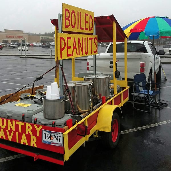 A red and yellow boiled peanuts trailer with large pots, propane tanks, and a colorful umbrella.