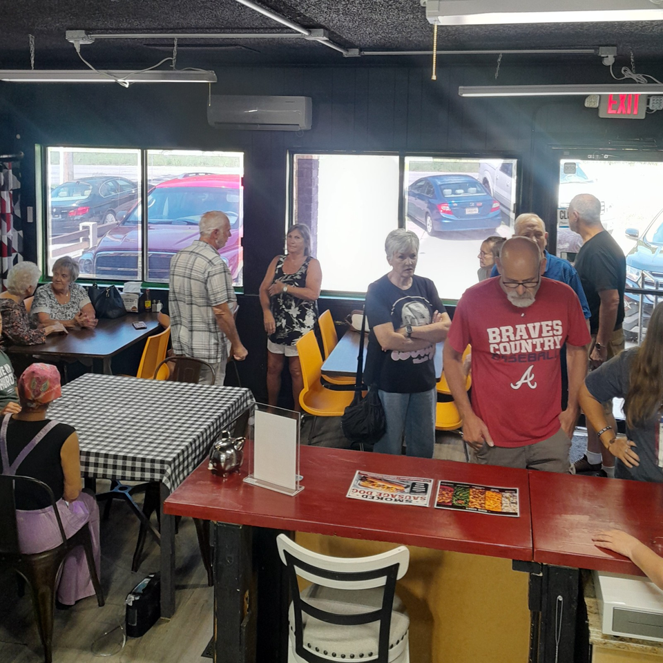 Busy casual restaurant interior with diners gathered around tables, checkered tablecloths, colorful chairs, and large windows.