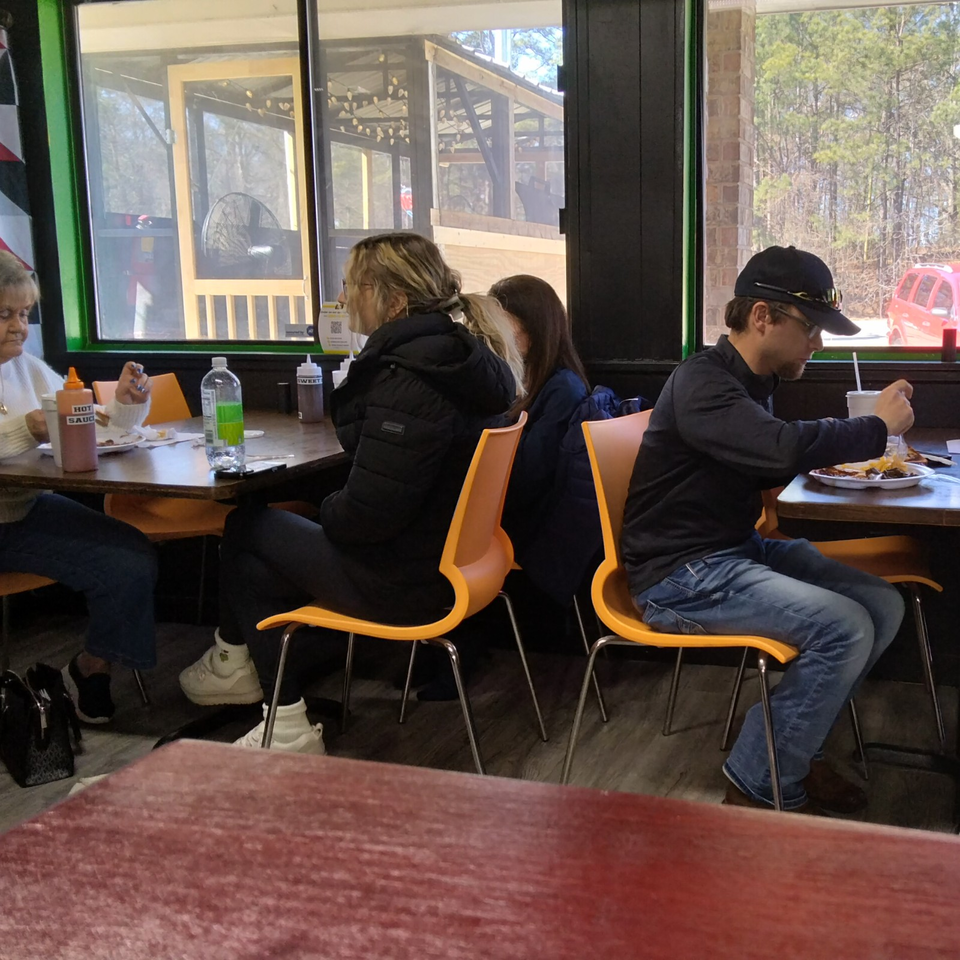 Diners sit at tables inside a casual restaurant, with hot sauce, drinks, and plates of food near large windows.