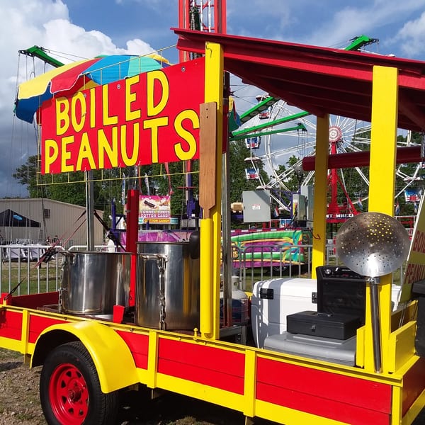 A red and yellow "Boiled Peanuts" stand with large cooking pots, at a fairground with a Ferris wheel.